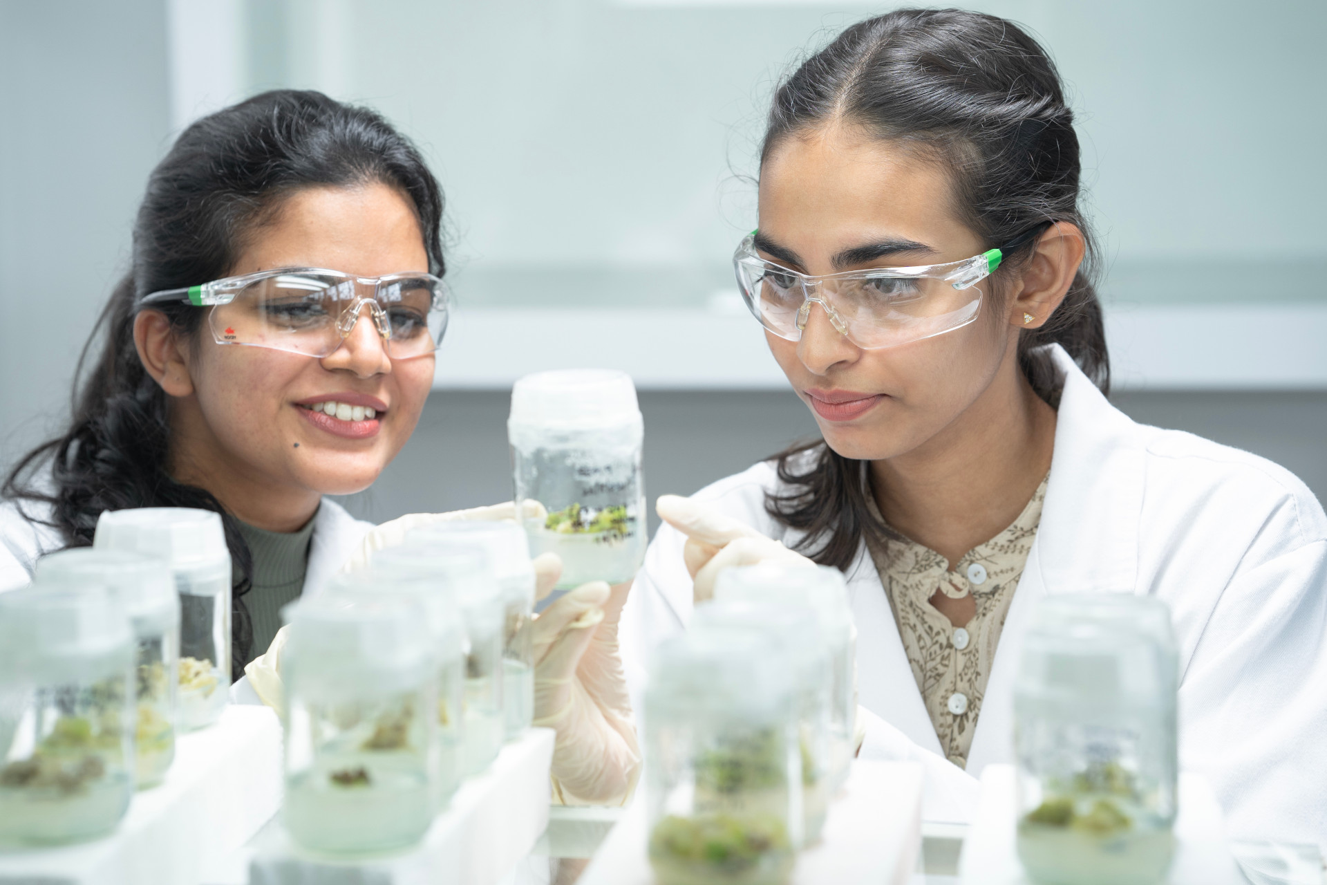 Two female researchers wearing lab coats and safety glasses looking at a beaker with content inside.